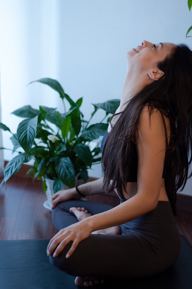 image shows a woman doing yoga at home she is dressed in black yoga clothes in an easy crossed legged position blog post is about practice yoga at home for beginners