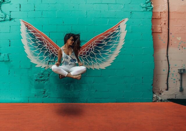 woman in easy crossed leg position flying up in the air in the background is a green wall with wings looks like her wings