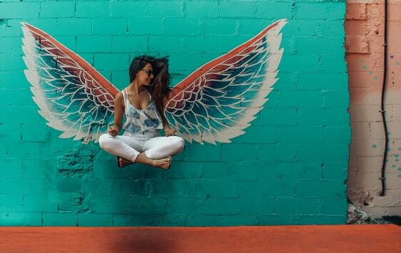 woman in easy crossed leg position flying up in the air in the background is a green wall with wings looks like her wings
