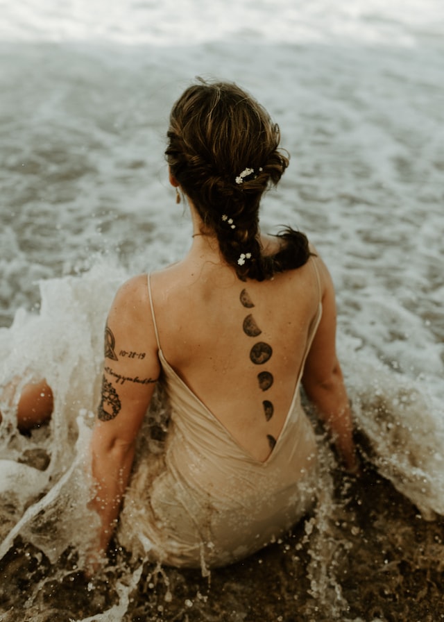 image showing woman sitting in moving water woman is wearing a light cream coloured dress and has moon phase tattoo on her back