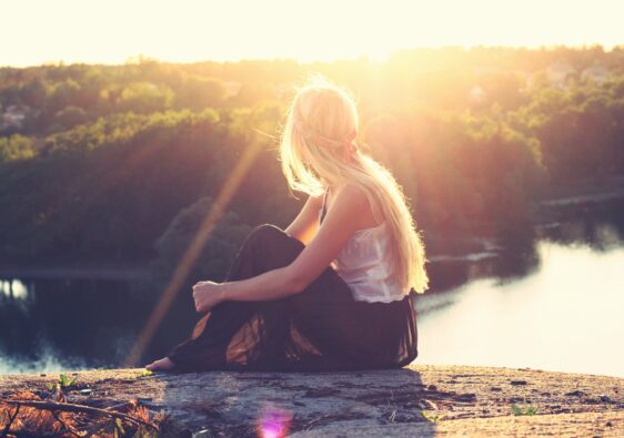 blonde girl sitting on top of mountain looking down at a lake evening in sunset