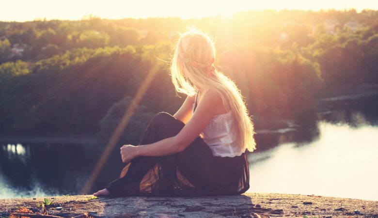 blonde girl sitting on top of mountain looking down at a lake evening in sunset