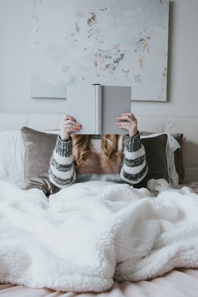 the picture shows a woman sitting in bed holding a book in front of her face and she is reading the blog post is about winter self-care ideas