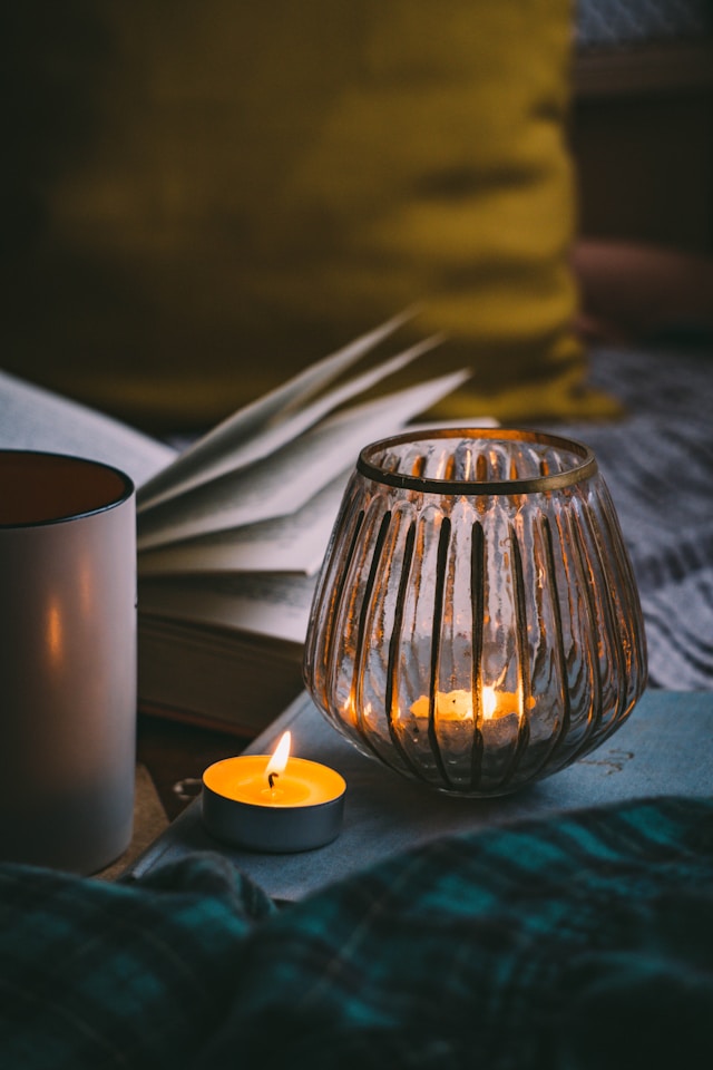 the picture shows a cozy indoor setting a close up of a tea light, a coffee cup and an open book; the blog post is about winter self-care ideas