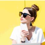 woman wearing big black sunglasses standing in front of a yellow wall holding a mobile phone how to practice self-care at work