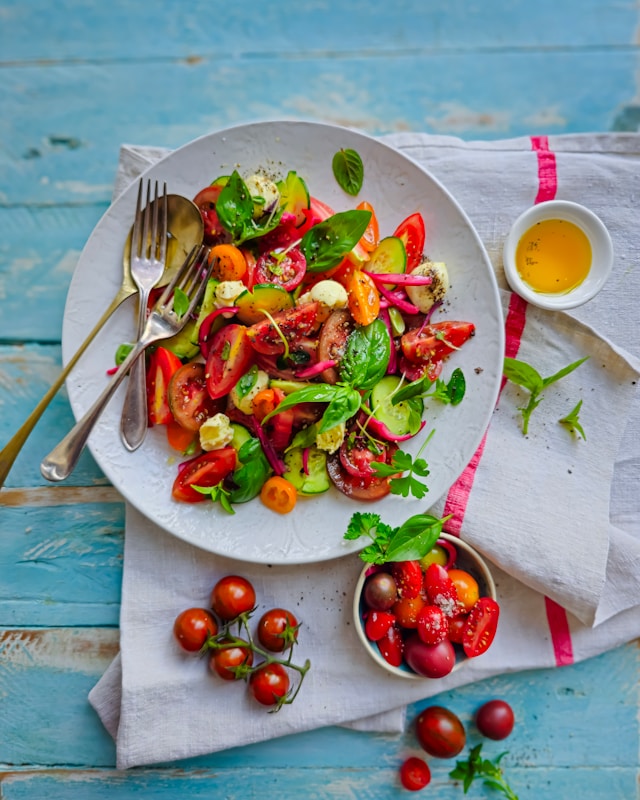 the picture shows a fresh summer salad placed on a white plate and a rustic looking light blue table surrounded by fresh tomatoes and salad dressing the blog post is about summer self-care ideas