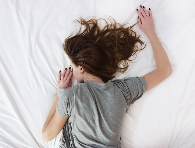woman with brown hair lying in bed on stomach wearing grey t-shirt sleeping feel good activities