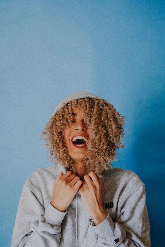 image showing woman with curly hair wearing a grey hoodie standing in front of a blue background woman is smiling and happy blog post is about feel good activities