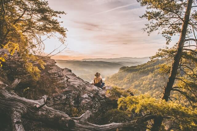 woman with long blonde hair and cowboy hat sitting within nature setting in mountains breathing in the air enjoying a break 11 easy ways to relax when stressed