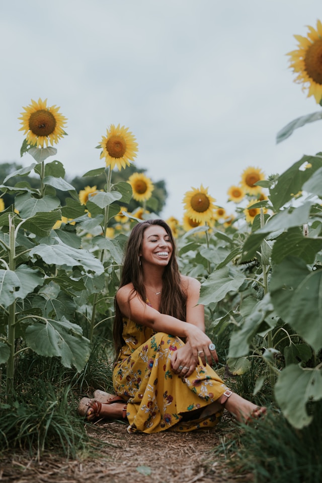 image showing a woman with long dark hair wearing a yellow dress sitting in sun flower field smiling blog post is about mindful glow up tips