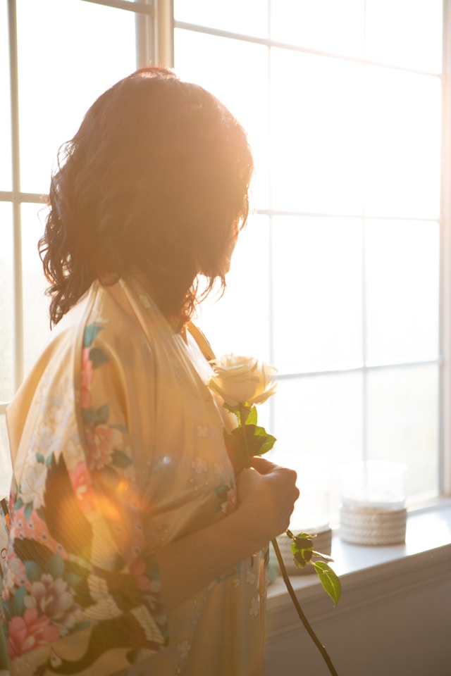 image shows a woman standing at a window holding a single white rose to her heart blog post is about self-love exercises