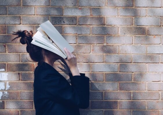 woman walking in front of brick wall holding a book to cover her face 7 ways to overcome procrastination