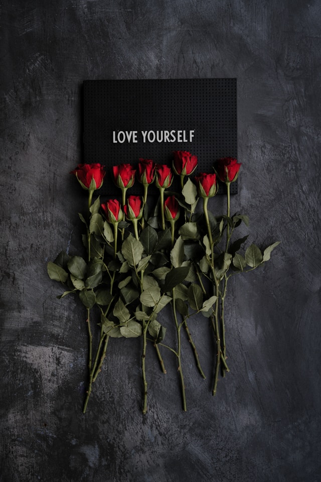 image features a black background and on the black background there is a sign saying love yourself in white lettering as well as red roses shown underneath the sign blog post is about affirmations for self love