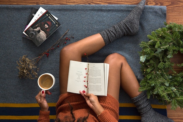picture shows a close up of women's legs woman wearing knitted long sweater and thick knitted socks holding a cup of tea in her left hand and a book in her right hand sitting on the floor self care products for busy women