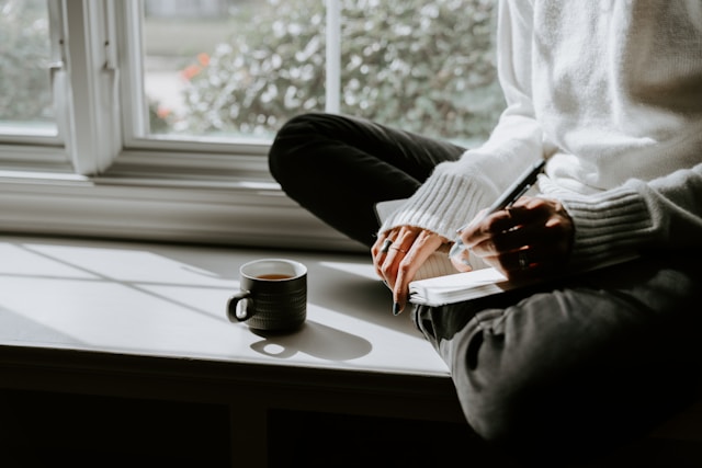 image shows a close up of a woman sitting on a window seal with her journal and a cup of tea blog post is about self reflection journal prompts for the end of the year