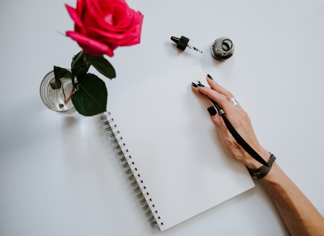 image shows a red rose next to a journal and a close up of a female hand with an ink pen blog post is about self reflection journal prompts for the end of the year