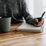 image shows a close up of a woman writing into a journal sitting at a wooden table with a cup of coffee or tea; the blog post is about personal development journal prompts for a mid year check in
