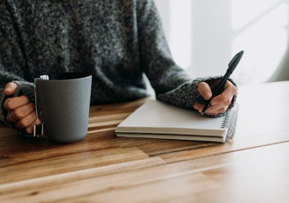 image shows a close up of a woman writing into a journal sitting at a wooden table with a cup of coffee or tea; the blog post is about personal development journal prompts for a mid year check in
