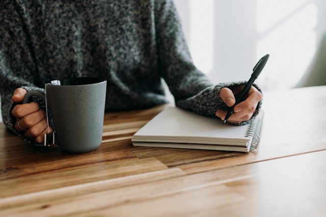 image shows a close up of a woman writing into a journal sitting at a wooden table with a cup of coffee or tea; the blog post is about personal development journal prompts for a mid year check in