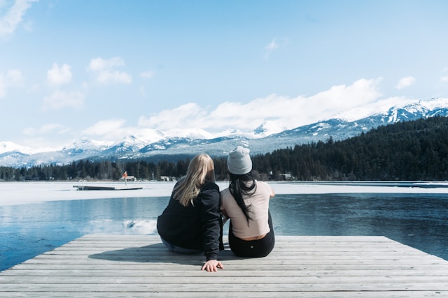 the picture shows two women from the back sitting on a wooden pier looking at a lake with mountains in the background the blog post is about how to simplify life and things to quit
